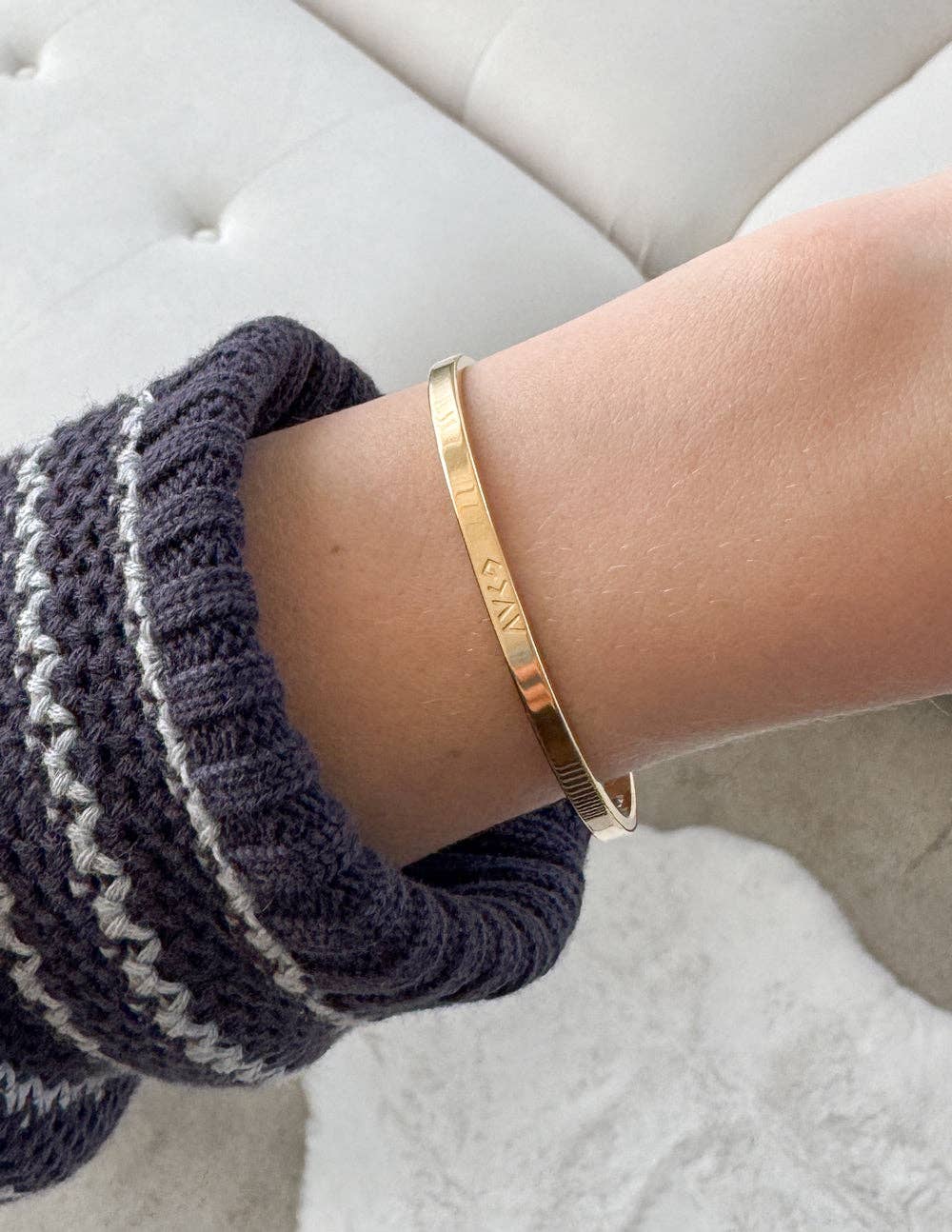 Gold bracelet on a wrist with a textured sleeve against a neutral background